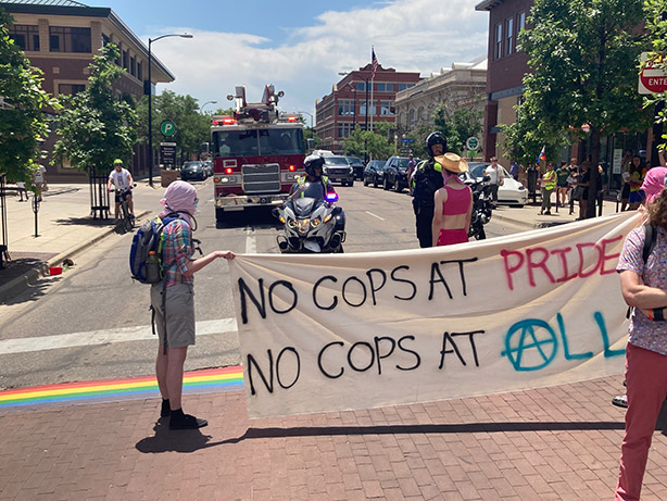 People holding a banner that says “No cops at pride / no cops at all” with two motorcycle cops behind it, presumably telling the demonstrators to move out of the way. A fire truck (the head of the pride motorcade) is behind them too
